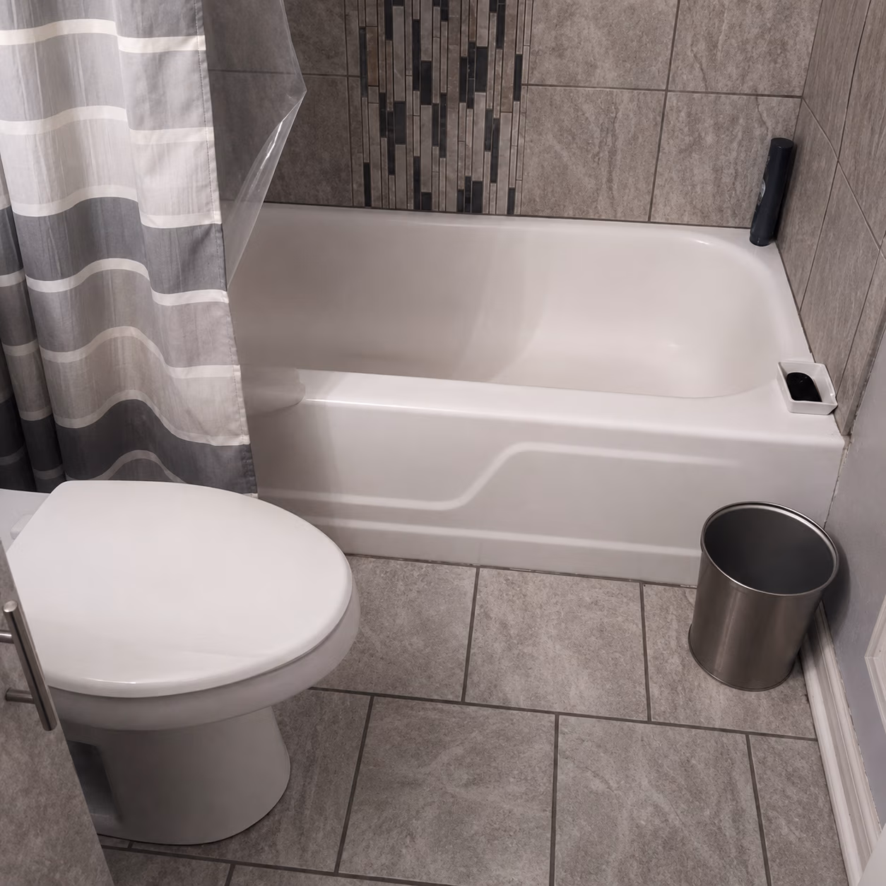 Bathroom with white toilet, bathtub, gray tiled floor and wall, striped shower curtain, and silver trash can.