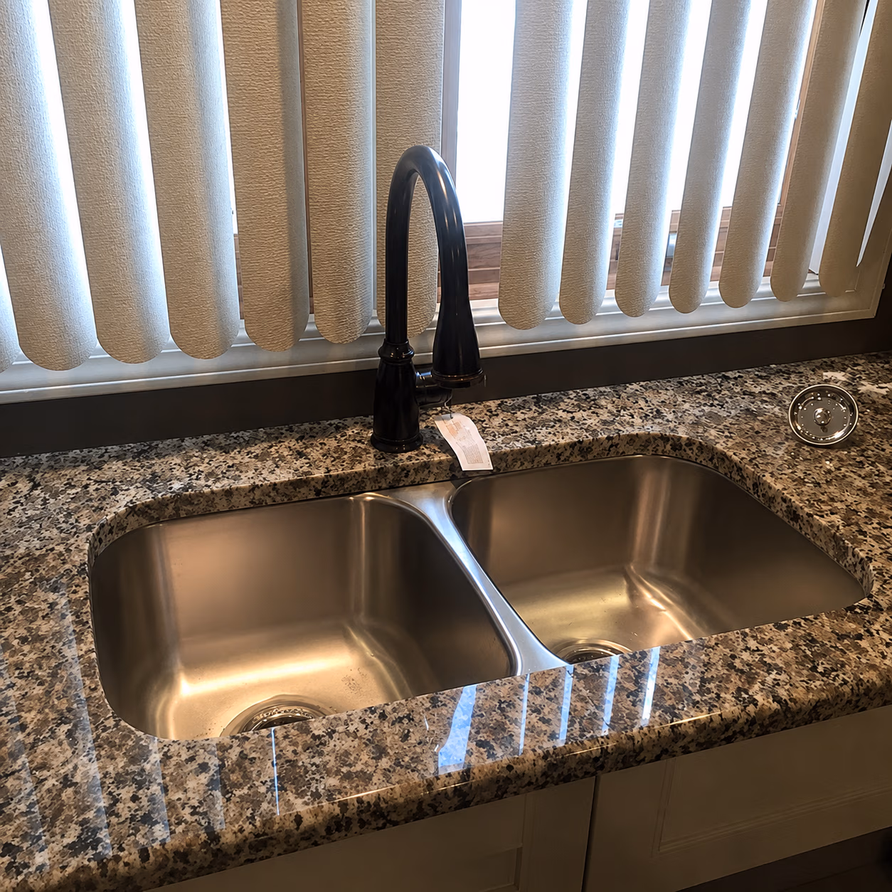 Double stainless steel kitchen sink with black curved faucet on granite countertop under window blinds.