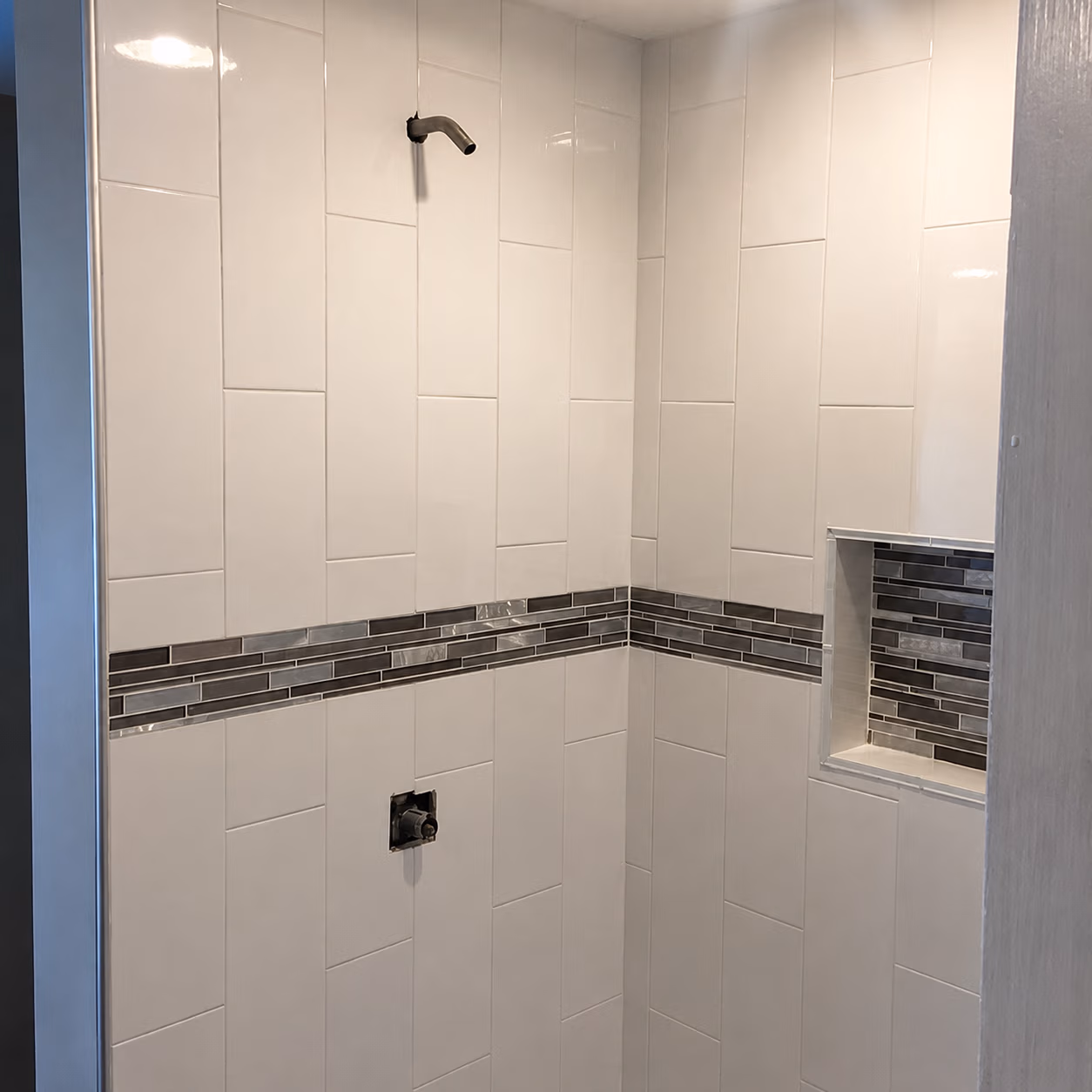 Tiled shower corner with white tiles, dark glass tile accent strip, and built-in niche.