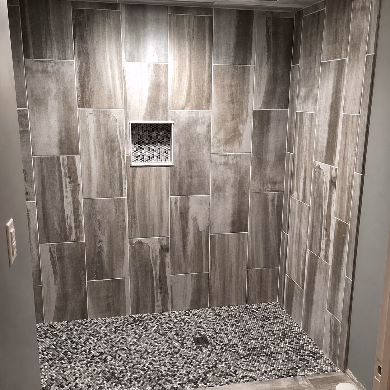 Walk-in shower with large gray tiles on walls and small pebble-patterned tiles on floor.