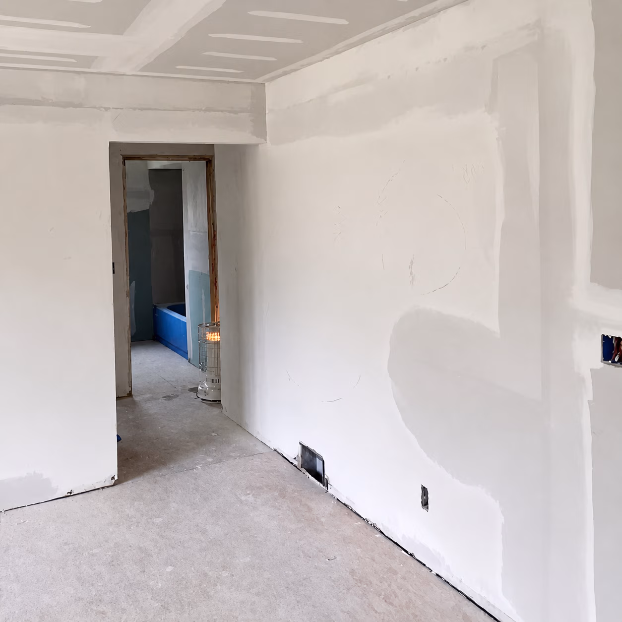 Room under construction with unfinished drywall and a heater near a doorway to a bathroom with a blue bathtub.