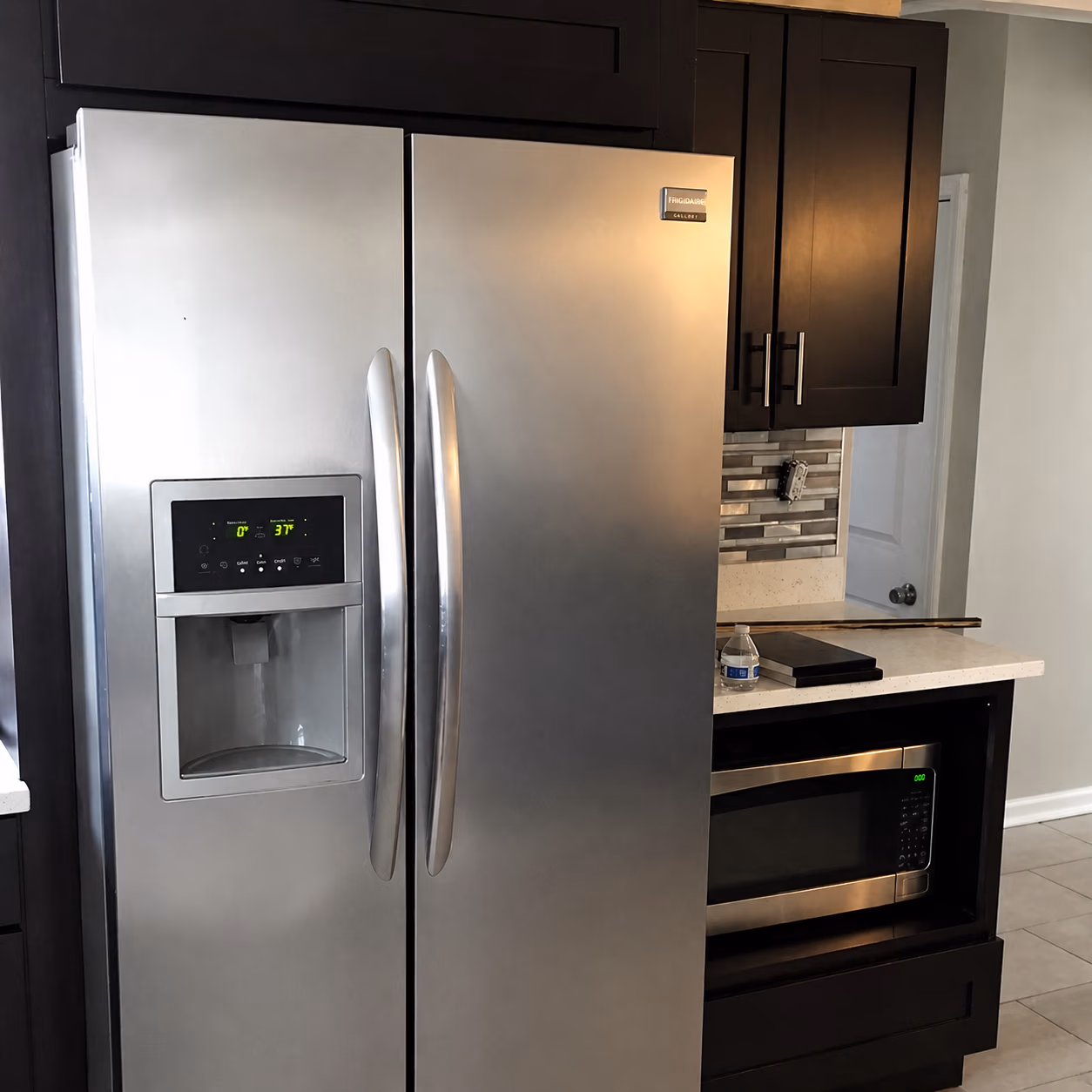Modern stainless steel fridge with water dispenser next to dark cabinets and built-in microwave.