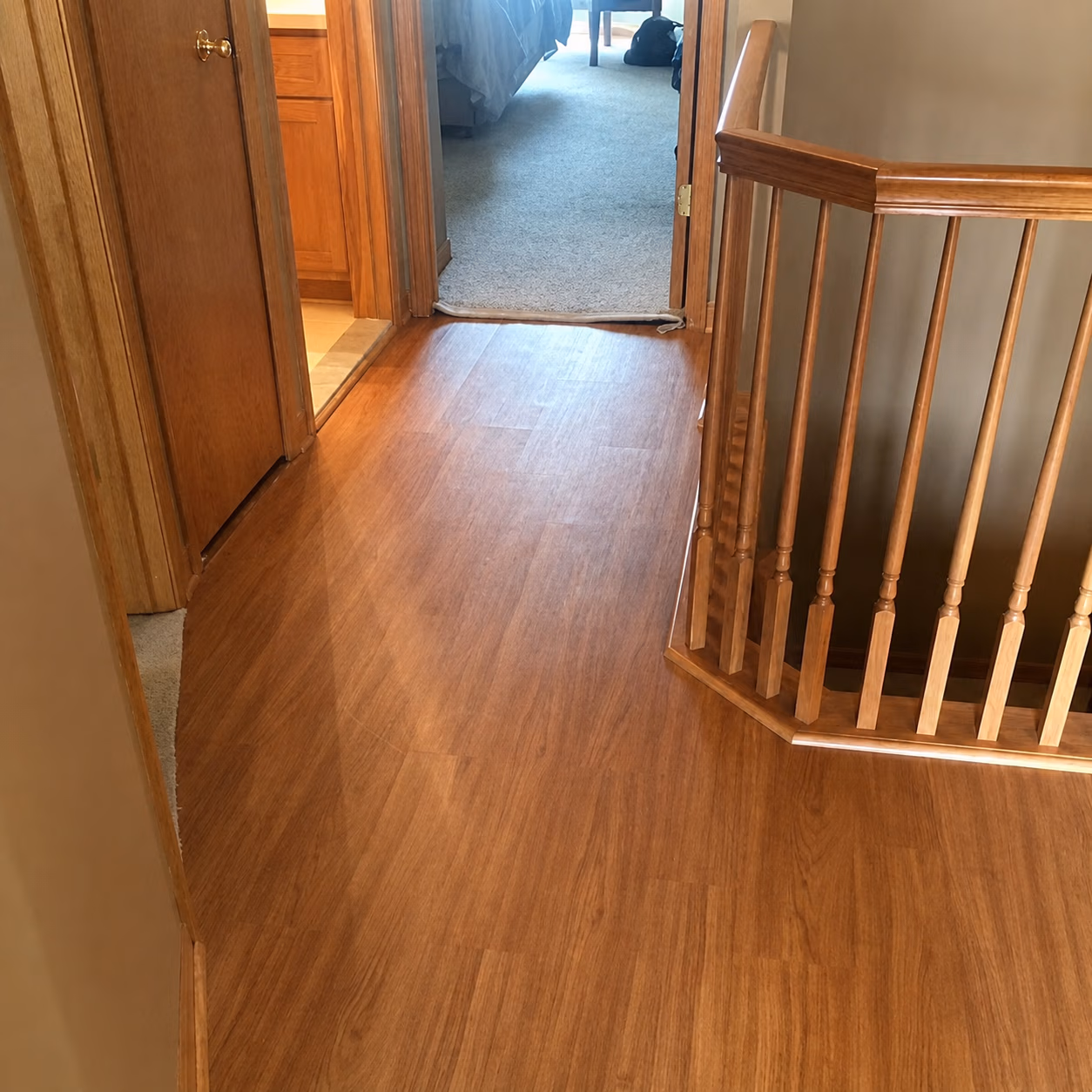 Indoor hallway with wood floor, wooden railing on right, and open door showing carpeted room ahead.