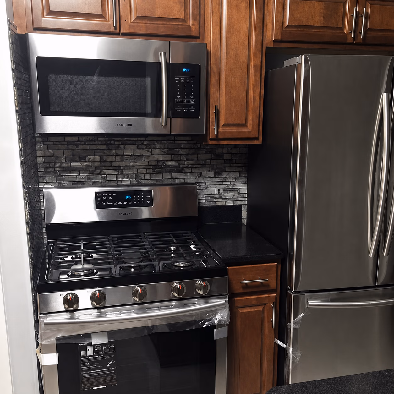 Modern kitchen section with stainless steel Samsung microwave, gas stove, and refrigerator beside wooden cabinets.