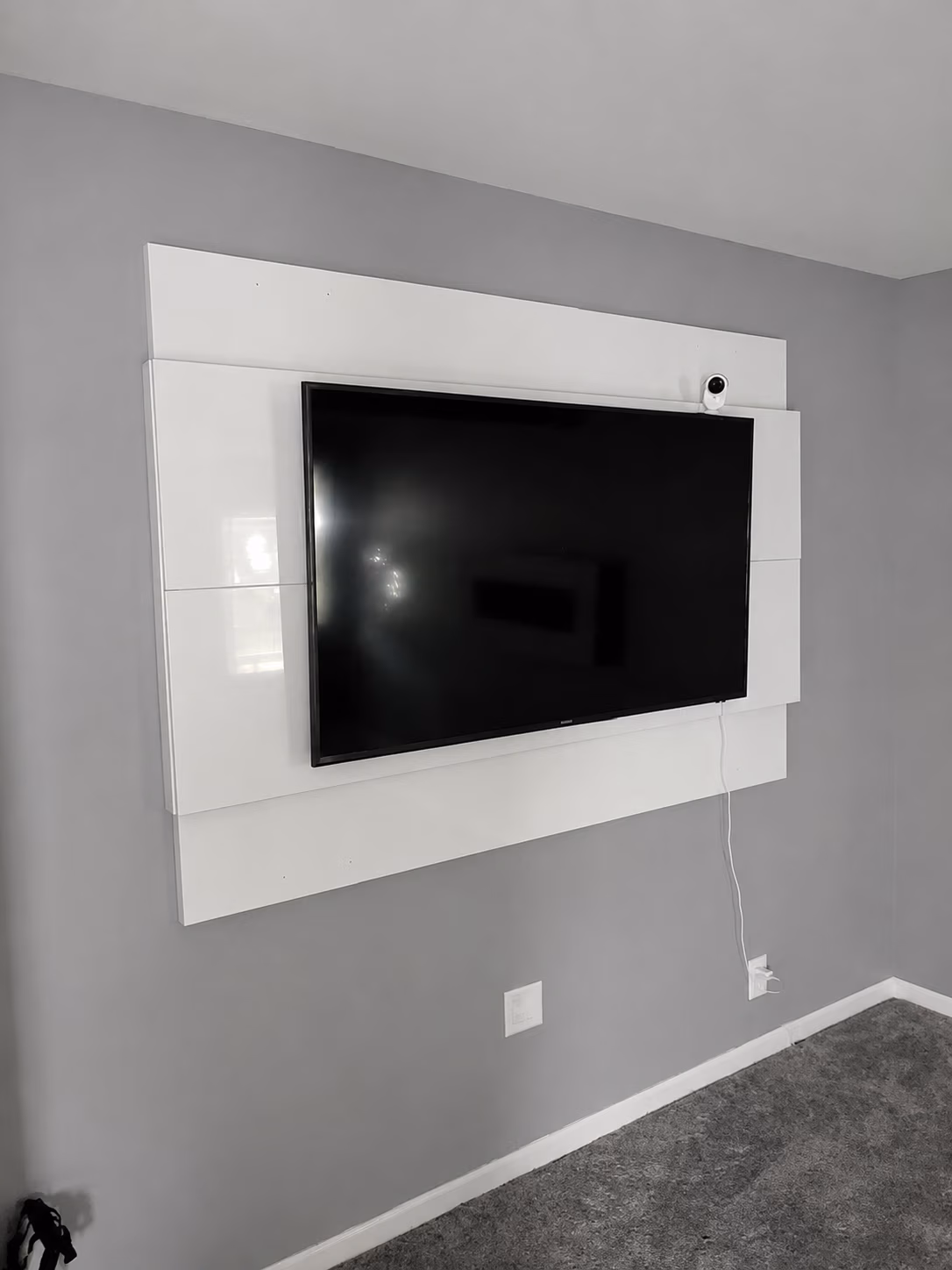 Flat screen TV mounted on a white panel against a gray wall with a small security camera above it.