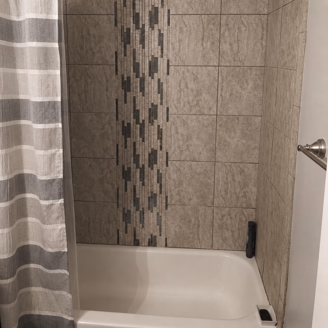 Empty bathtub with gray tiled walls and a striped gray and white shower curtain.