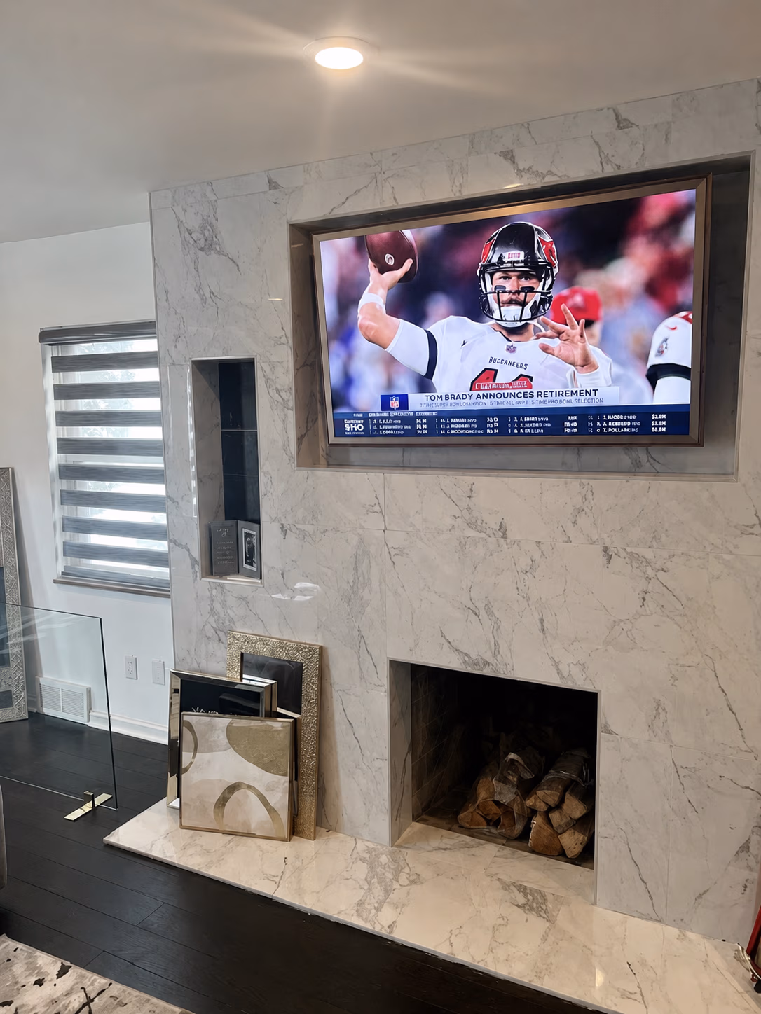 Modern marble fireplace with stacked wood and TV showing Tom Brady announcing retirement.