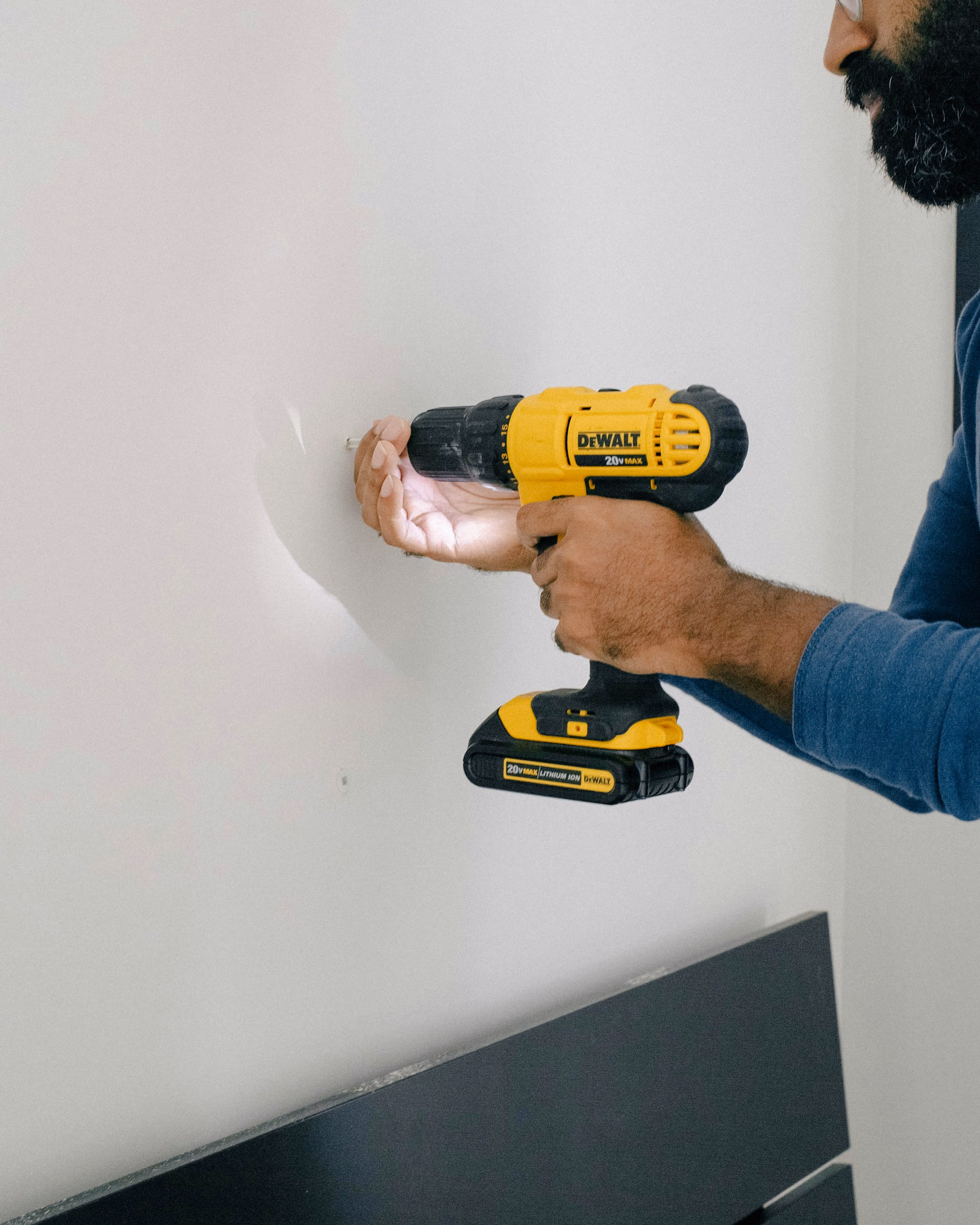 Person using a yellow DeWalt cordless drill to drill into a white wall indoors.
